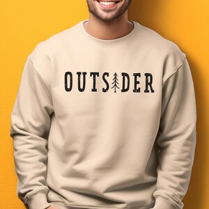 May include: A beige crewneck sweatshirt featuring the black text "OUTSIDER" printed across the chest. The letter "I" is replaced by a stylized black pine tree graphic. The lettering has a distressed, textured appearance throughout the design.