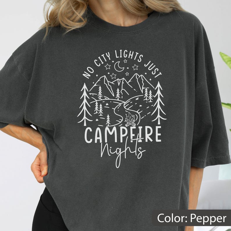 May include: A charcoal grey t-shirt featuring a white graphic print of a mountain landscape with pine trees, a crescent moon, stars, and a campfire. The text reads NO CITY LIGHTS JUST CAMPFIRE Nights in a mix of block and script lettering.
