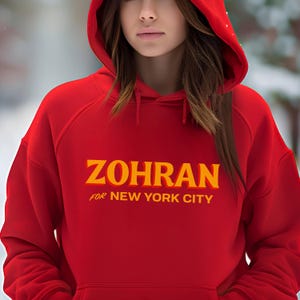 May include: A red hooded sweatshirt with the word "ZOHRAN" in yellow block letters, with "FOR NEW YORK CITY" below. The sweatshirt has a drawstring hood and a front pocket.