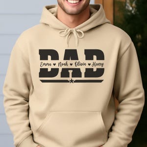 May include: A tan hoodie with a black graphic that says "DAD" in bold letters. Below the word "DAD" are the names Emma, Noah, Olivia, and Henry, each separated by a heart symbol. A star is below the names.
