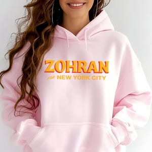 May include: A light pink hoodie with the word "ZOHRAN" in orange and yellow lettering, with the text "for NEW YORK CITY" underneath. The hoodie has a drawstring hood and a front pocket.