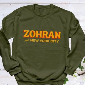 May include: Olive green sweatshirt with the name "ZOHRAN" in orange block letters with a red outline. Below the name, the text "FOR NEW YORK CITY" is printed in orange. The sweatshirt is a crew neck style.