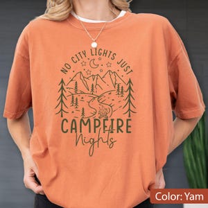 May include: A burnt orange t-shirt featuring a dark green graphic of a mountain landscape, pine trees, a campfire, a crescent moon, and stars. The text reads NO CITY LIGHTS JUST CAMPFIRE Nights in a mix of block and script lettering.