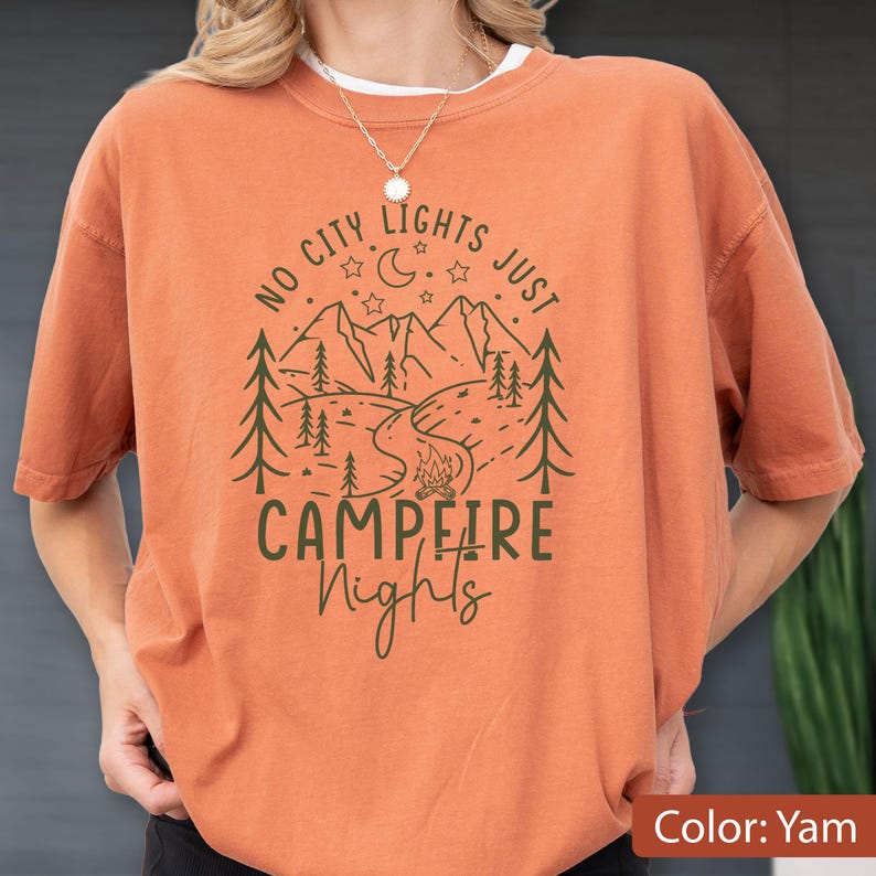 May include: A burnt orange t-shirt featuring a dark green graphic of a mountain landscape, pine trees, a campfire, a crescent moon, and stars. The text reads NO CITY LIGHTS JUST CAMPFIRE Nights in a mix of block and script lettering.