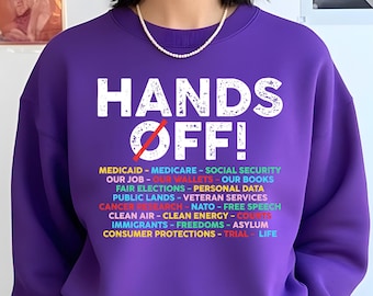 May include: Purple sweatshirt with the text "HANDS OFF!" in white with a red slash through the O in OFF. Below the text is a list of items in various colors: MEDICAID, MEDICARE, SOCIAL SECURITY, OUR JOB, OUR WALLETS, OUR BOOKS, FAIR ELECTIONS, PERSONAL DATA, PUBLIC LANDS, VETERAN SERVICES, CANCER RESEARCH, NATO, FREE SPEECH, CLEAN AIR, CLEAN ENERGY, COURTS, IMMIGRANTS, FREEDOMS, ASYLUM, CONSUMER PROTECTIONS, TRIAL, LIFE.
