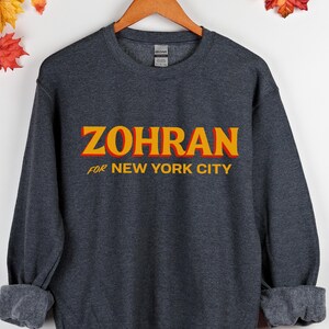 May include: A dark gray sweatshirt with the word "ZOHRAN" in large, orange and yellow letters across the chest. Below the word "ZOHRAN" is the text "FOR NEW YORK CITY" in smaller, white letters. The sweatshirt is hanging on a wooden hanger.