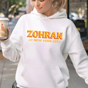 May include: White hoodie with the word "ZOHRAN" in orange and yellow lettering, with "FOR NEW YORK CITY" underneath. The hoodie has a front pocket and a hood. The person in the image is holding a coffee cup.
