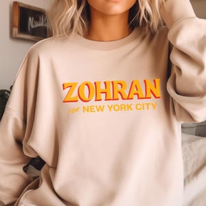 May include: A beige sweatshirt with the word "ZOHRAN" in orange and yellow block letters with a red shadow. Below the word is the phrase "FOR NEW YORK CITY" in smaller orange letters. The sweatshirt is a neutral color.
