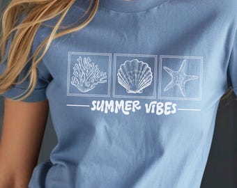 May include: A blue jean-coloured t-shirt with a white graphic of a coral, seashell, and starfish in square boxes. The words "SUMMER VIBES" are printed below. The shirt has short sleeves.