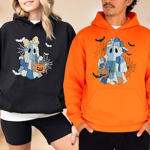 May include: A black hoodie and an orange hoodie, each featuring a graphic of a ghost made of blue and tan patchwork fabric. The ghosts hold orange jack-o'-lantern buckets, surrounded by black spiderwebs and bats. The black hoodie ghost wears a bow.