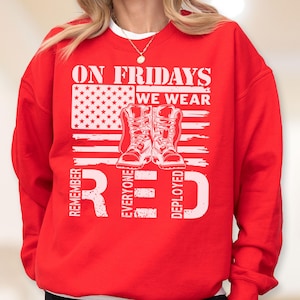 May include: A bright red crewneck sweatshirt featuring a white graphic print. The design includes the text ON FRIDAYS WE WEAR, a stylized American flag, a pair of military boots, and the text REMEMBER EVERYONE DEPLOYED in large white lettering.