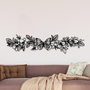 以下が含まれることがあります： 蝶と様々な花や葉をあしらった黒い金属製のフローラルウォールアート。装飾品は、枕と毛布が付いた茶色のソファの上にある白い壁に取り付けられています。アートワークは水平デザインです。