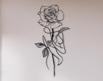 May include: Black metal wall art featuring a stylized rose with a human figure incorporated into the stem. The art piece is mounted on a white wall in a modern interior setting.