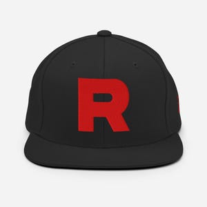 May include: A black snapback cap featuring a large, bold, red letter R embroidered on the front centre. The hat has a structured crown, a flat brim, and a small red detail visible on the side panel, maintaining a clean and simple design.