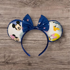 May include: Blue headband with a blue bow and white fabric ears featuring Mickey Mouse, Minnie Mouse, Donald Duck, and Daisy Duck characters.