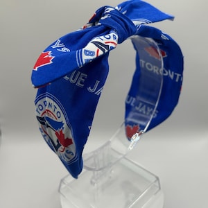 May include: A blue fabric headband with a knotted bow, displaying the Toronto Blue Jays logo and text. The headband is designed to be worn on the head. The fabric is patterned with the team's logo.
