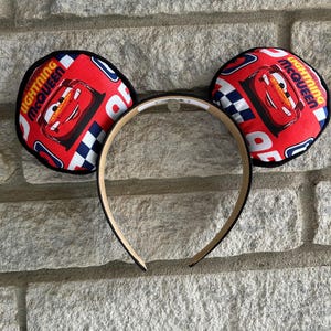 May include: Red and black mouse ears with a Lightning McQueen image on the front. The ears are made of fabric and have a black headband.