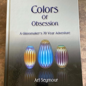 Puede incluir: Un libro titulado "Colors of Obsession" con el subtítulo "A Glassmaker's 70 Year Adventure". La portada presenta tres piezas de arte en vidrio coloridas y estampadas en tonos naranja, azul y morado. El nombre del autor, Art Seymour, está en la parte inferior.