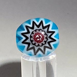 Könnte beinhalten: Eine runde, blaue Glasperle mit einem Sternmuster. Die Perle hat einen roten Mittelkreis mit den Buchstaben "AS" und "MM" darin. Das Sternmuster ist schwarz und weiß, mit einem hellblauen Hintergrund.