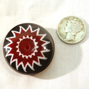 May include: A round, black glass paperweight with a red and white starburst design. A silver coin is next to the paperweight for size comparison. The paperweight has a glossy finish and a unique, eye-catching pattern.