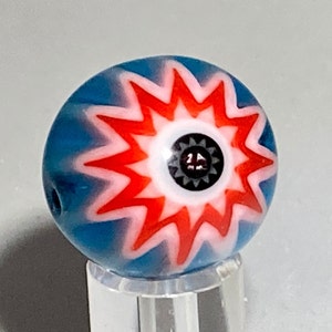 Könnte beinhalten: Eine runde Glasperle mit blauer Grundfarbe und einem rot-weißen Sternmuster. Die Mitte der Perle zeigt ein kleines schwarz-weißes augenähnliches Muster. Die Perle hat ein Loch zum Auffädeln.