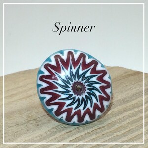 May include: A round glass bead featuring a central hole and a radial pattern of alternating maroon, white, and teal zig-zag lines that spiral outward from the center. The word Spinner is printed in black text above the bead.