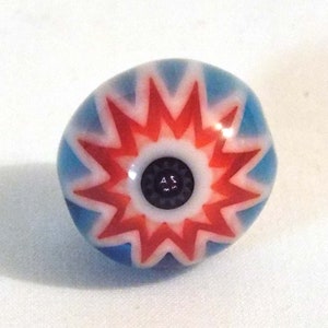 Könnte beinhalten: Eine runde Glasperle in Blau, Rot und Weiß mit einem Sternmuster. Die Mitte ist schwarz mit weißem Text. Diese Perle ist wahrscheinlich für Schmuck- oder Bastelprojekte gedacht.