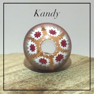 Könnte beinhalten: Eine runde, durchscheinende bernsteinfarbene Glasperle mit einem Mittelloch und einem dekorativen Millefiori-Muster aus weißen und roten sternförmigen Blumenmotiven. Das Wort Kandy ist in schwarzer Schrift über der Perle gedruckt.