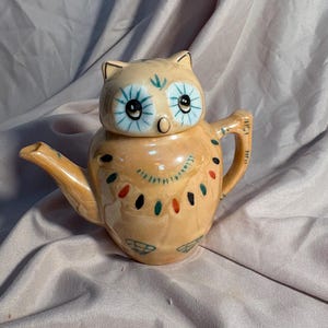 May include: A ceramic owl-shaped teapot in a light orange color. The teapot features a lid that forms the owl's head, with blue eyes and a beak. The body is decorated with colorful dots and lines. The teapot has a handle and a spout.