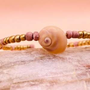 May include: A pink and gold beaded bracelet with a seashell charm. The seashell is a light brown color with a pink swirl in the center.