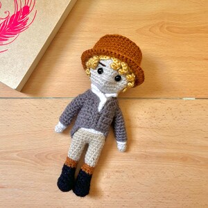 May include: A crocheted doll with brown hair, wearing a brown hat, a gray jacket, white shirt, brown pants, and black boots.