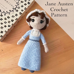 Puede incluir: Muñeca de ganchillo, inspirada en Jane Austen, con un vestido azul claro, un gorro blanco y pelo castaño. La muñeca tiene un cuello blanco y un cinturón azul. El texto "Jane Austen Crochet Pattern" es visible.