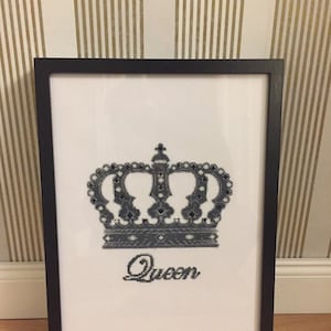 May include: Black and white cross-stitch embroidery in a black frame. The embroidery depicts a crown with jewels and the word "Queen".