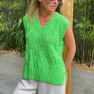 Może przedstawiać: Jasnozielona, bezrękawnikowa kamizelka z dekoltem w serek i wzorem w kratę. Kamizelka jest wykonana z dzianiny i ma prążkowany dół. Osoba nosi okulary przeciwsłoneczne i jasne spodnie.