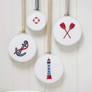 Puede incluir: Cuatro aros de bordado blancos con diseños náuticos rojos y azules. Los diseños incluyen una boya salvavidas, un faro, un ancla y un par de remos.