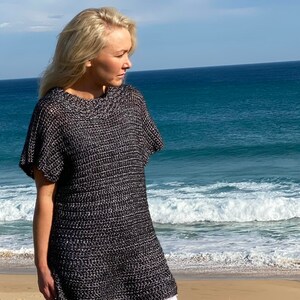 May include: A black and white knit tunic top with a loose fit and short sleeves. The model is wearing the top with white jeans and is standing on a beach.