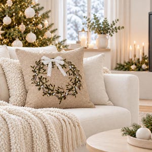 May include: A square throw cushion with a textured beige fabric cover featuring an embroidered green mistletoe wreath design centred on the front. A white fabric bow is attached at the top of the wreath, adding a decorative accent to the piece.