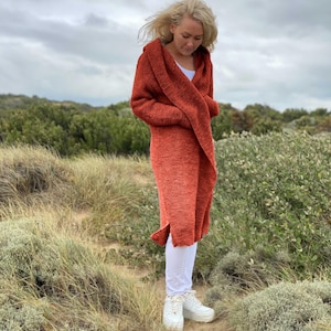 May include: A long, rust-coloured knitted cardigan with a hood. The cardigan is worn over white trousers and trainers. The person is standing in a field of tall grass and bushes under a cloudy sky. The cardigan has a ribbed texture.