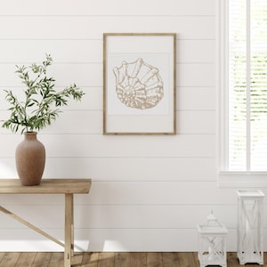 May include: A framed print of a seashell in a light brown color, mounted on a white background. The frame is a light wood color. The print is hanging on a white wall. A vase with greenery sits on a wooden table.