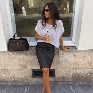 May include: A white short-sleeved knit top with a loose weave, a black knee-length skirt, a dark brown leather handbag with a buckle strap, and dark brown thong sandals. The outfit is paired with dark-tinted sunglasses and a gold chain necklace.
