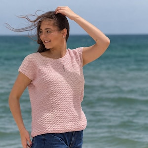 May include: A light pink short-sleeved top with a textured, woven knit pattern, a rounded neckline, and a straight hemline. The garment features a consistent, repeating stitch design throughout the front, providing a subtle, tactile surface finish.