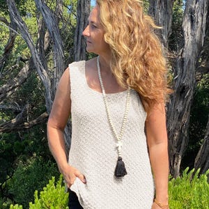 May include: A sleeveless beige knit tank top with a textured weave, paired with a long beaded necklace featuring light-colored wooden beads, a small white cross charm, and a dark brown tassel hanging at the center of the chest.