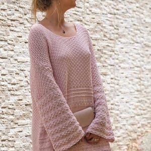 May include: A light pink, long-sleeved sweater dress with a textured knit pattern. The dress has a scoop neckline and flared sleeves. A small, light-colored clutch is held in front. The background is a light-colored stone wall.