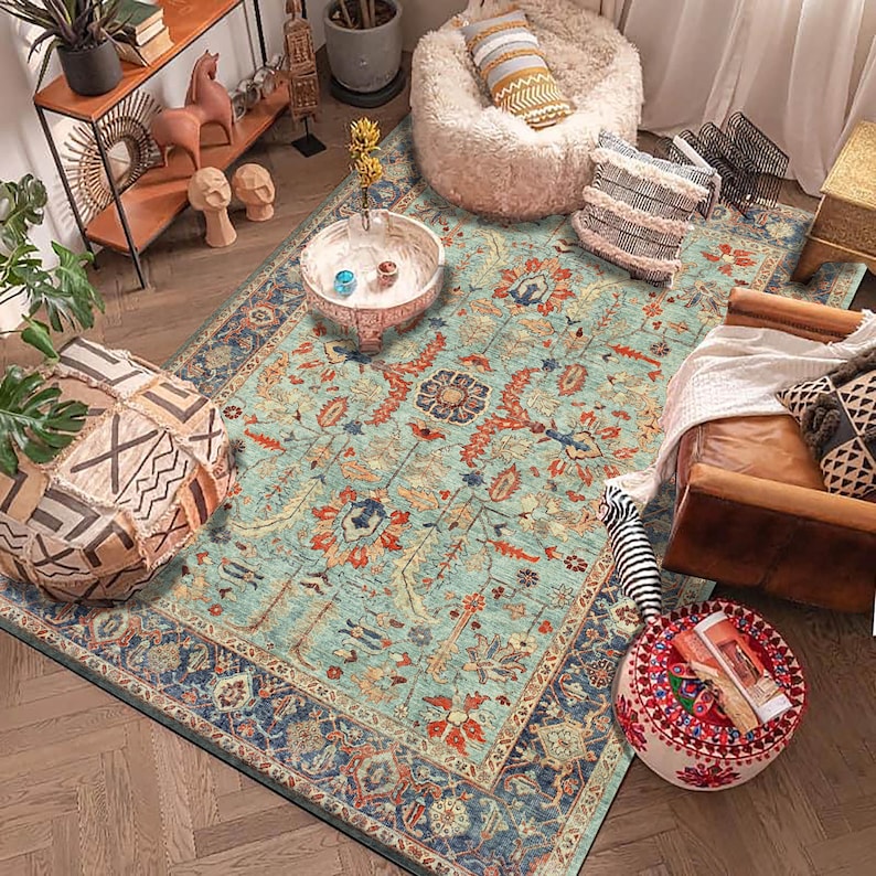 May include: A teal blue area rug with a floral pattern in shades of red, orange, and brown. The rug has a dark blue border with a geometric pattern. The rug is on a wooden floor and there are various decorative items in the background.