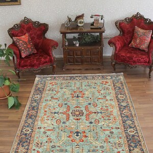 May include: A teal blue rug with a traditional Persian design. The rug features a central medallion with floral motifs and a border with intricate patterns. The rug is on a wooden floor in a room with two red velvet armchairs and a wooden table.