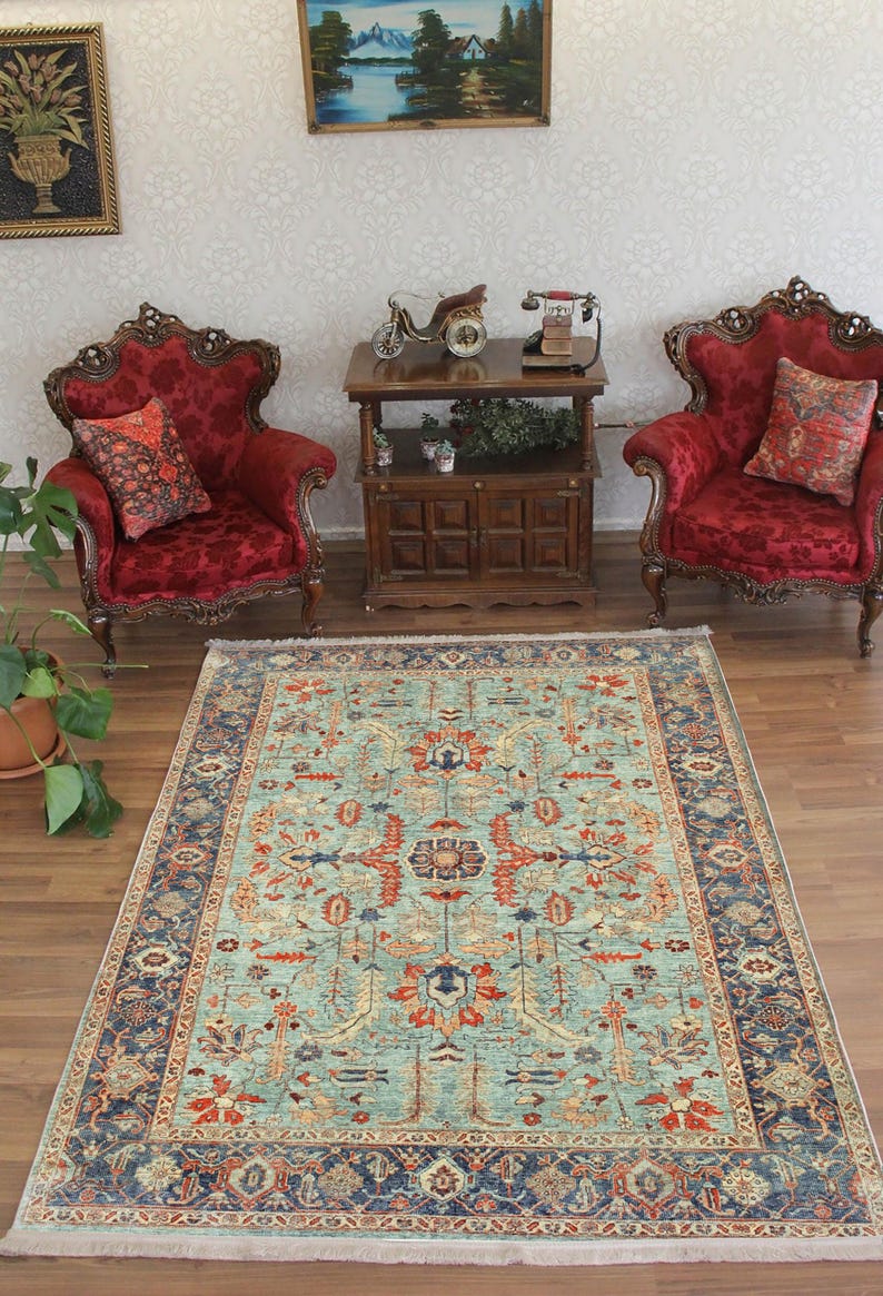 May include: A teal blue rug with a traditional Persian design. The rug features a central medallion with floral motifs and a border with intricate patterns. The rug is on a wooden floor in a room with two red velvet armchairs and a wooden table.