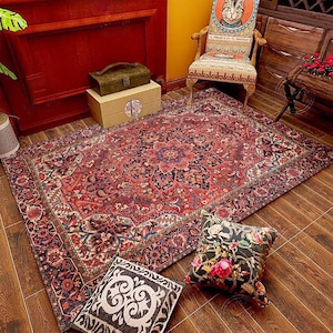 May include: A red, blue and cream patterned rug with a floral design. The rug is on a wooden floor with two throw pillows on top of it. One pillow is black with white floral designs and the other is grey with a swirl pattern.