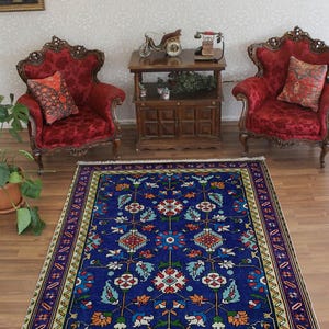 May include: A blue and white patterned rug with a floral design. The rug has a cream-coloured border with a geometric pattern. The rug is on a wooden floor in a room with two red velvet chairs and a wooden table.