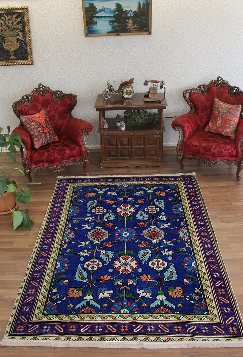 May include: A blue and white patterned rug with a floral design. The rug has a cream-coloured border with a geometric pattern. The rug is on a wooden floor in a room with two red velvet chairs and a wooden table.