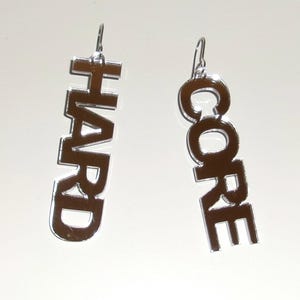 Könnte beinhalten: Ein Paar hängende Ohrringe aus verspiegeltem silberfarbenem Acryl. Ein Ohrring zeigt das Wort HARD in einer vertikalen Blockschrift, der andere das Wort CORE in einer vertikalen Blockschrift. Beide Ohrringe sind an silberfarbenen Metallhaken befestigt.